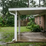 Rain running off a white aluminum patio cover into a seamless gutter, discharging through a single downspout onto a splash block away from the slab, at a brick home in south Louisiana