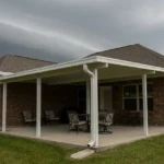 Brick home with a white aluminum patio cover and seamless gutters under a dark squall line, ground still dry