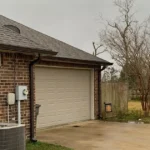 Dark seamless gutter and downspout on a red brick home beside the garage, with bare deciduous trees past the driveway