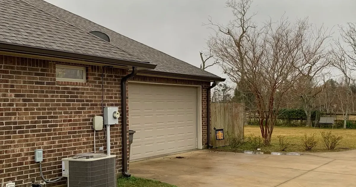 Dark seamless gutter and downspout on a red brick home beside the garage, with bare deciduous trees past the driveway