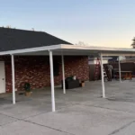 White aluminum patio cover going up over a wide concrete slab behind a brick home, with ladders and a crew member at the far end