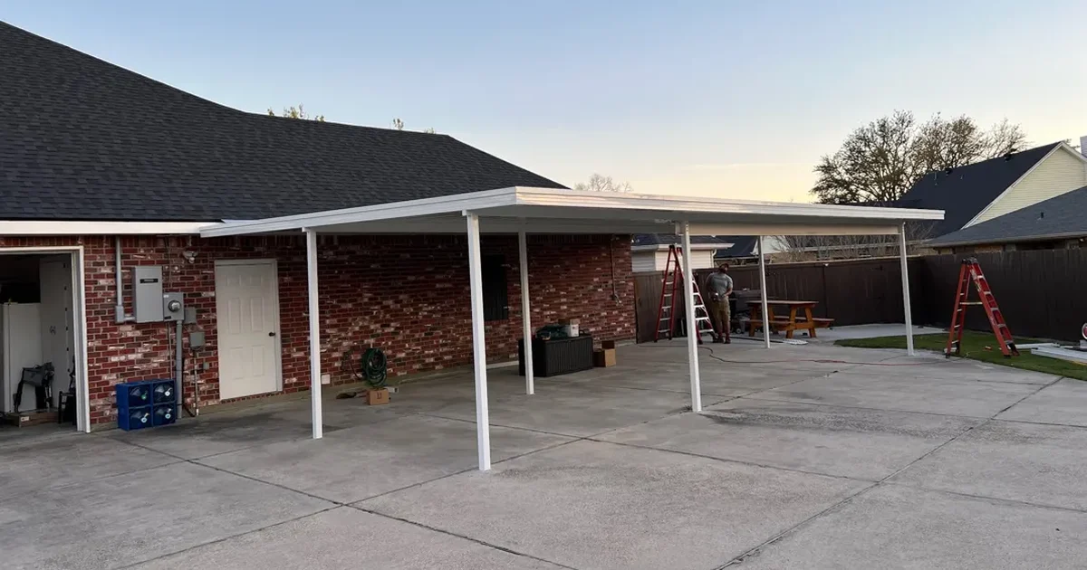 White aluminum patio cover going up over a wide concrete slab behind a brick home, with ladders and a crew member at the far end