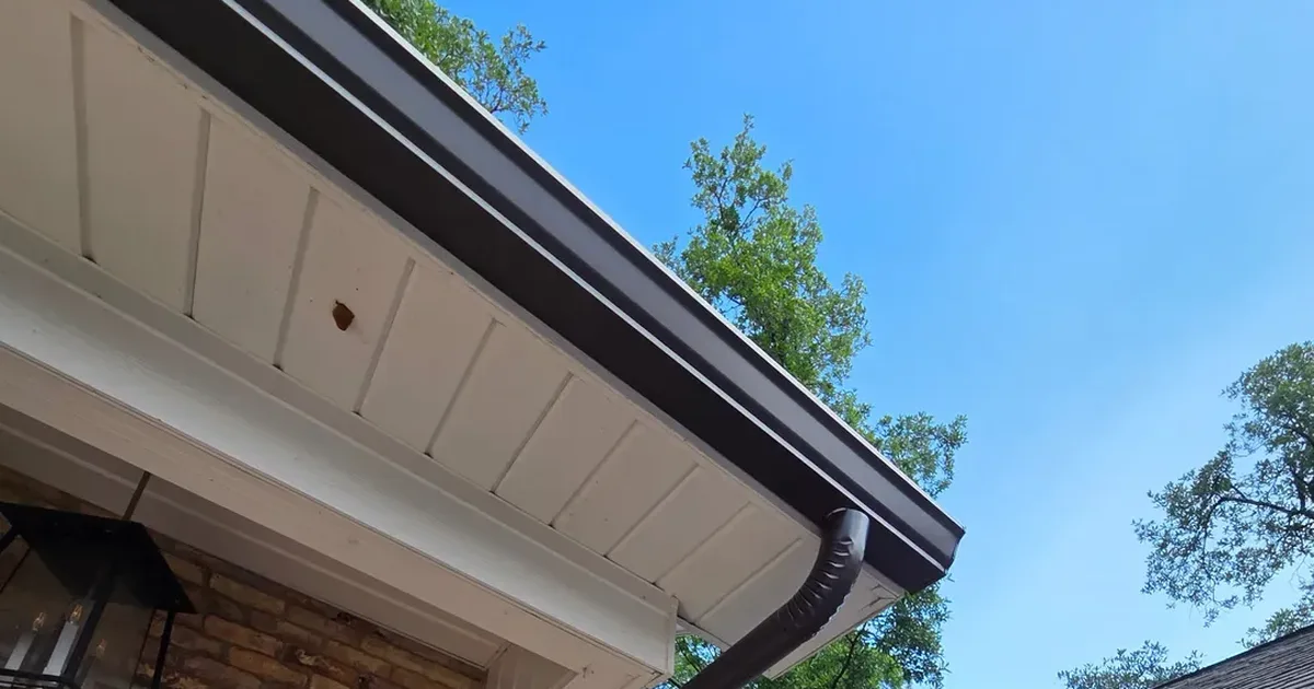 Brown seamless gutter and downspout on a brick home with live oak branches above the roofline