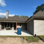 New concrete patio behind a Livingston Parish area home, with fresh sod and bare soil in the yard and a downspout drain line running away from the house