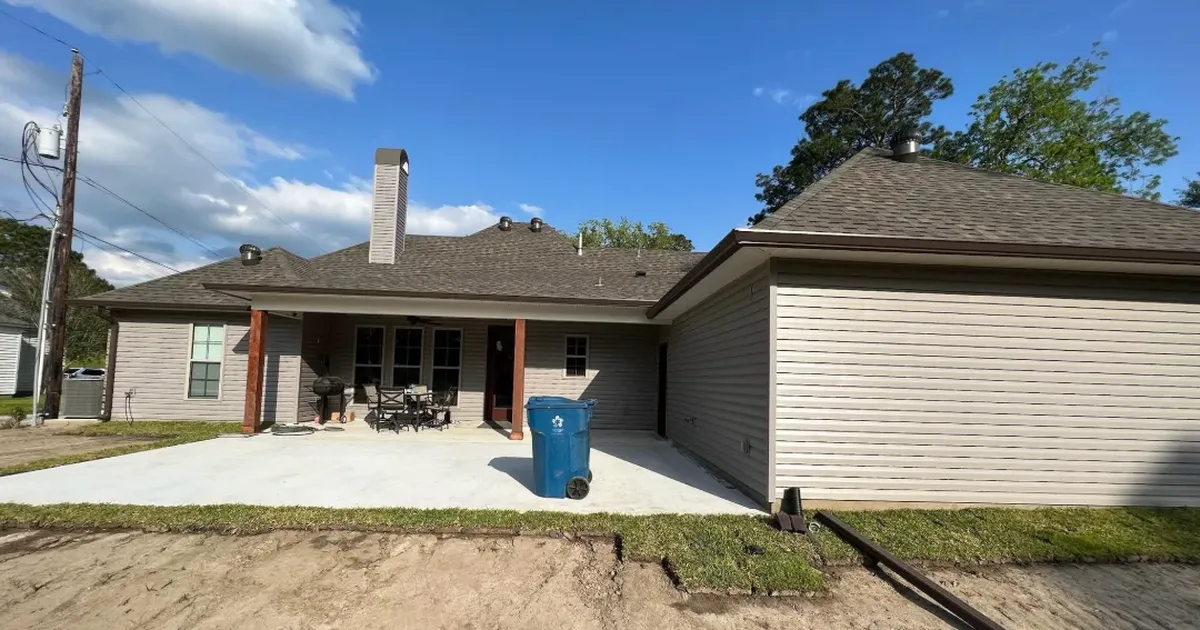 New concrete patio behind a Livingston Parish area home, with fresh sod and bare soil in the yard and a downspout drain line running away from the house