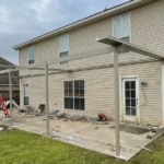 Patio cover frame going up over a concrete patio behind a two-story home, with roof panels laid out on the lawn