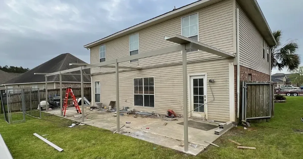 Patio cover frame going up over a concrete patio behind a two-story home, with roof panels laid out on the lawn