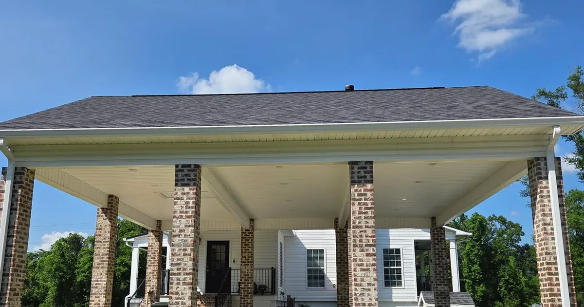 Long white seamless gutter running the full front edge of a wide brick-column carport, with a downspout at each end